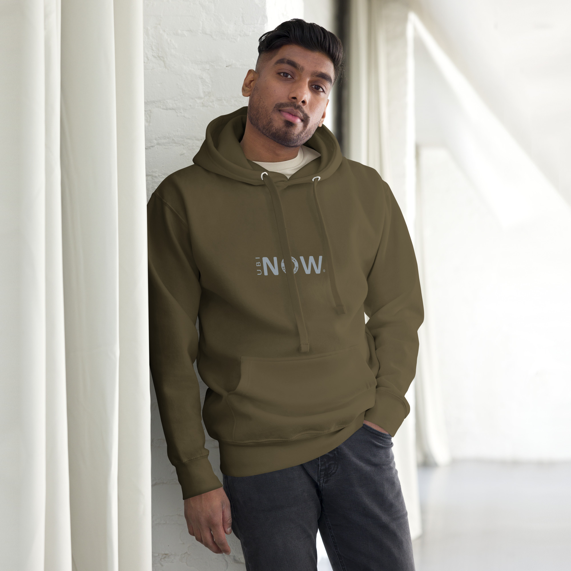 Hombre con sudadera con capucha color oliva apoyado en una pared blanca.